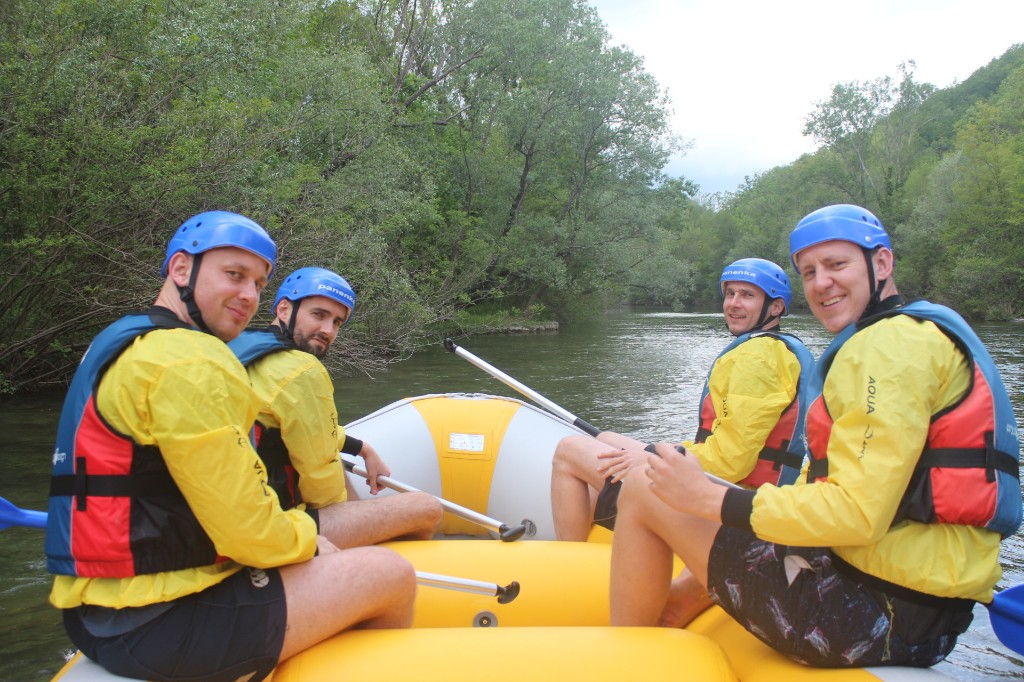 Four rafters in helmets and yellow jackets in a yellow inflatable raft on the river with green trees in the background